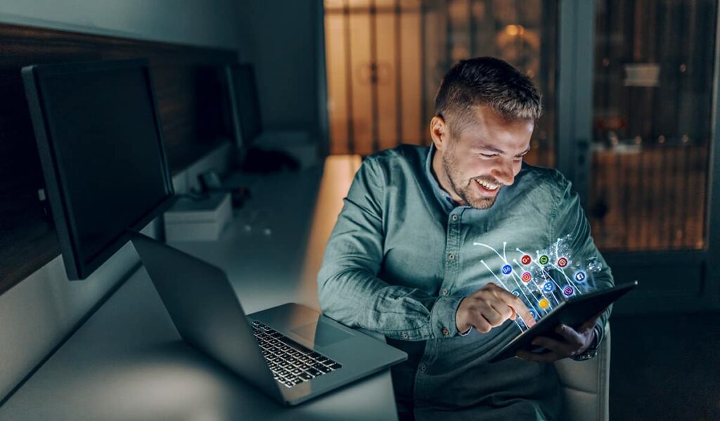 Handsome Caucasian Bearded Freelancer With Toothy Smile Sitting In Office Late At Night And Using Tablet For Surfing The Internet Social Media Concept